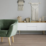 lichte beige eiken vloer met brede planken in rechte vloerpatroon onder een witte modern design dressoir met houten blad, groene fluwelen fauteuil en wanddecoratie van macramé en keramische accessoires