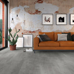 Donkergrijze tegelvloer met brede stroken en matte structuur in een moderne woonkamer met bakstenen muur, oranje stoffen bank en plantenpot met cactus.