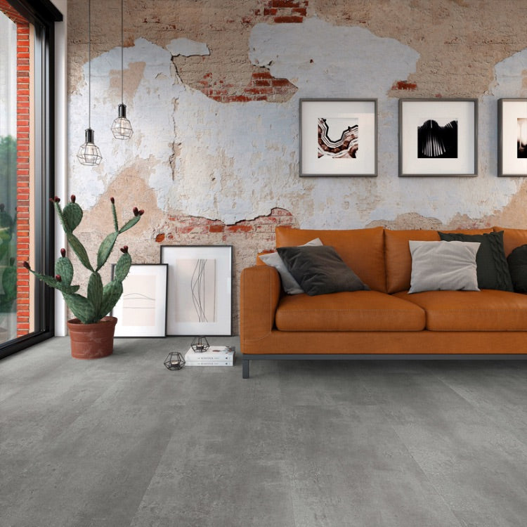 Donkergrijze tegelvloer met brede stroken en matte structuur in een moderne woonkamer met bakstenen muur, oranje stoffen bank en plantenpot met cactus.