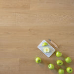 lichte eiken vloer met brede planken en subtiele houtstructuur, daarop een wit marmeren snijplank met een gesneden groene appel en een klein mes met houten handvat, omringd door meerdere hele groene appels