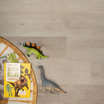 licht beige eiken vloer met brede planken en subtiele houtstructuur, hoek van een rond bruin-beige kleed met witte accenten en twee speelgoed dinosaurussen naast een felgeel kinderboek over dinosaurussen