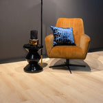 licht eiken vloer met brede planken en subtiel houtnerfstructuur, moderne cognacbruine fauteuil met zwarte metalen voet en blauw fluwelen kussen met crocopatroon, zwart glazen bijzettafeltje met gouden accent
