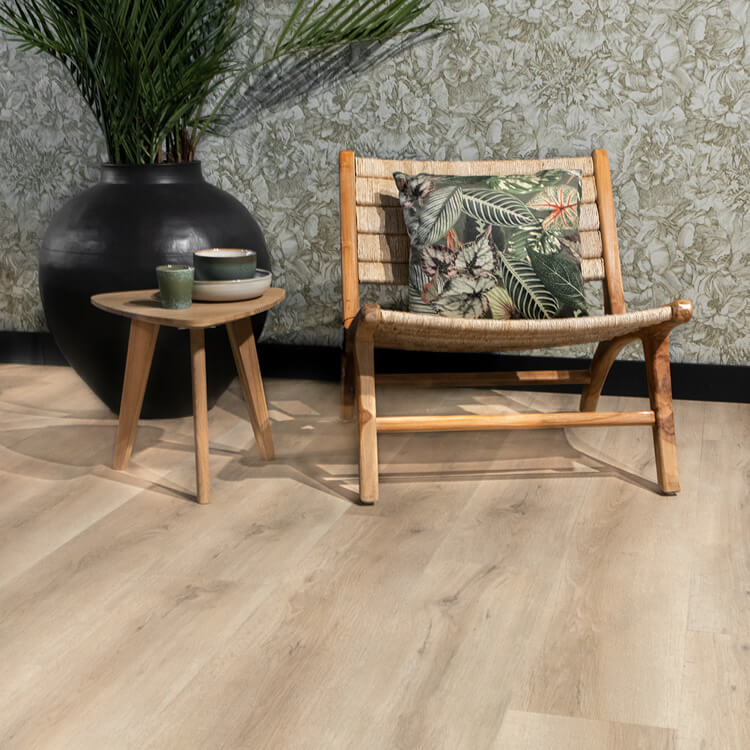 lichte eiken vloer met brede planken en een subtiele houtnerf structuur, gecombineerd met een laag houten bijzettafeltje, een gevlochten houten stoel met een kussen met tropisch bladmotief en een grote matzwarte vaas met groene palmbladplant, tegen een muur met een groen-grijze botanische behangprint