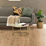 licht beige eiken vloer met natuurlijke houtstructuur en zichtbare noesten, moderne grijs stoffen bank met decoratieve kussens, ronde glazen salontafel met goudkleurig metalen frame, plantenmand met groene kamerplant, neutrale beige wandkleur