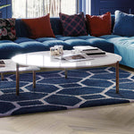Lichte eiken visgraatvloer met matte afwerking onder een donkerblauw vloerkleed met wit geometrisch patroon en een witte ronde salontafel met metalen poten.