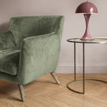 licht eiken visgraatvloer met brede planken, groene fluwelen fauteuil met schuine houten poten en ronde glazen bijzettafel met gouden frame en roze designlamp