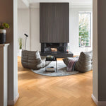 Visgraatvloer met brede planken in licht eiken houtkleur met subtiele houtnerfstructuur, gelegen in een woonkamer met moderne zithoek en ronde grijs-beige vloerkleed.