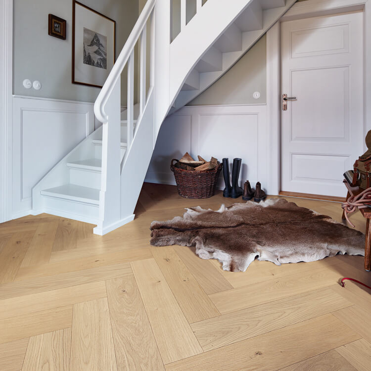 lichte eiken vloer in visgraatpatroon met brede planken, natuurlijke houtstructuur en warme beige tint, in een entree met witte trap, witte deur en bruine dierenhuid als vloerkleed