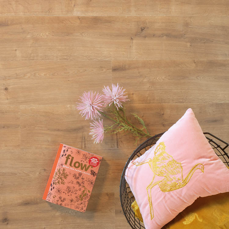 lichte beige eiken vloer met subtiele houtnerfstructuur, zachte roze bloemen en een roze kussen met geel geborduurde vogel in een zwart metalen mand