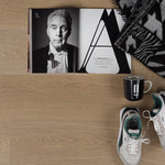 lichte laminaatvloer in eikenstructuur met fijne houtnerf en subtiele beige tint, met opengevouwen zwart-wit magazine, zwart-witte geprint deken, zwarte mok met witte tekst en wit-groene sneakers op de vloer geplaatst