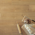 licht bruin eiken vloer in brede planken met subtiele nerfstructuur, op de vloer een houten deegroller en een snijplank met gesneden volkorenbrood, bloem en een mes