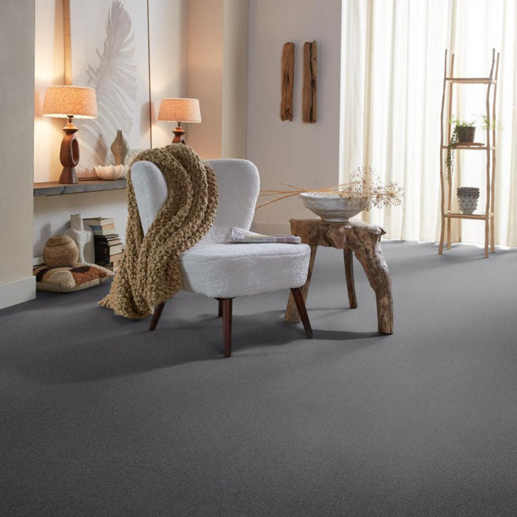 Grijs tapijt met fijne structuur in een moderne woonkamer, gecombineerd met een witte fauteuil met houten poten en een grove, beige gebreide deken.