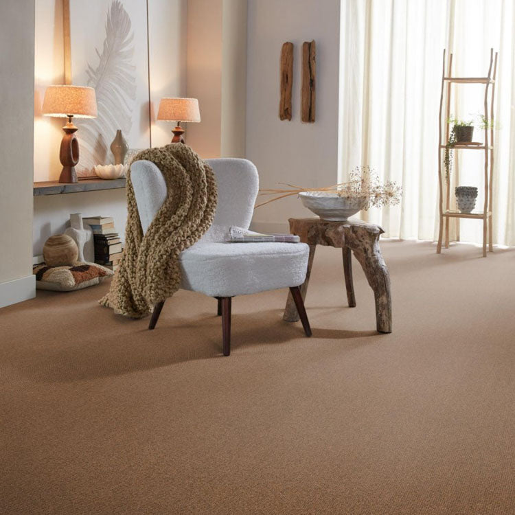 bruin tapijt met fijne, gelijkmatige structuur in een warme, natuurlijke kleur, gecombineerd met een witgrijze fauteuil met zachte stof en een grof, bruin gebreid plaid in een lichte woonkamer met zachte, neutrale verlichting