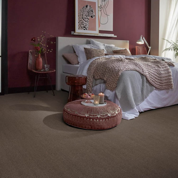 slaapkamer met taupe kleurig effen tapijt, decoratieve ronde poef met mandala patroon in oudroze en lichte plaids op het bed tegen een muur geschilderd in donker bordeaux rood