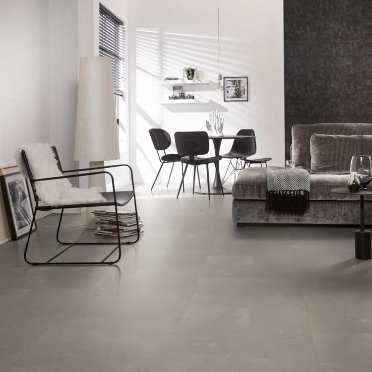 lichte vloer met grote vierkante tegels in mat asgrijs met subtiele golvende textuur, moderne zithoek met donkergrijze stoffen bank en zwarte minimalistische eetkamerstoelen aan ronde tafel in lichte ruimte met veel natuurlijk licht