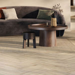 Licht beige visgraatvloer met subtiele houtstructuur en brede planken in een moderne woonkamer met ronde houten salontafel en donkergrijze gebogen bank.