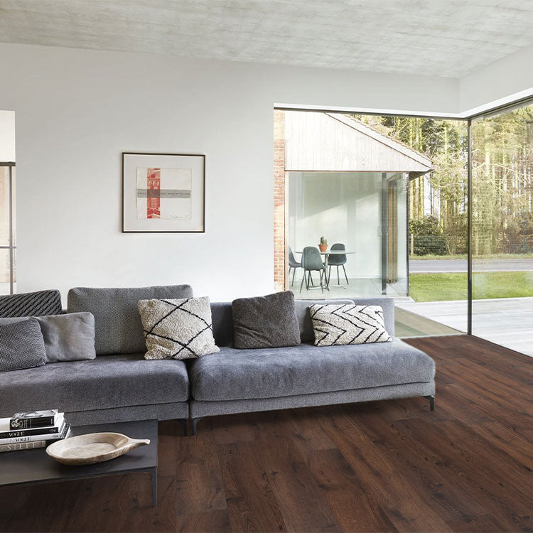 donkerbruin vloer met brede planken en lichte nerven in houtstructuur, moderne woonkamer met grote ramen en grijze stoffen bank met zwarte en beige kussens