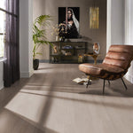 lichte vloer in licht grijs eiken kleur met subtiele houtnerfstructuur, brede planken in rechte vloerige legwijze in een modern interieur met leren fauteuil en open kast tegen taupe kleurige muur