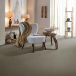 Zacht taupe kleurig tapijt met een egale, fijne structuur in een lichte, sfeervolle woonruimte met een moderne lichtgrijze fauteuil met bruine houten poten en een grove gebreide camel kleurige plaid.