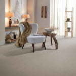 zachte effen beige tapijtvloer in een lichte woonkamer met een lichtgrijze fauteuil met houten poten en een grove bruin-beige gebreide plaid, natuurlijke houten bijzettafel en neutrale decoratie met zachte gordijnen voor veel lichtinval