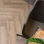Licht beige eiken vloer in visgraatpatroon met subtiele houtnerfstructuur, hoek van een grijze stoel en een gedeelte van een vloerkleed zichtbaar.