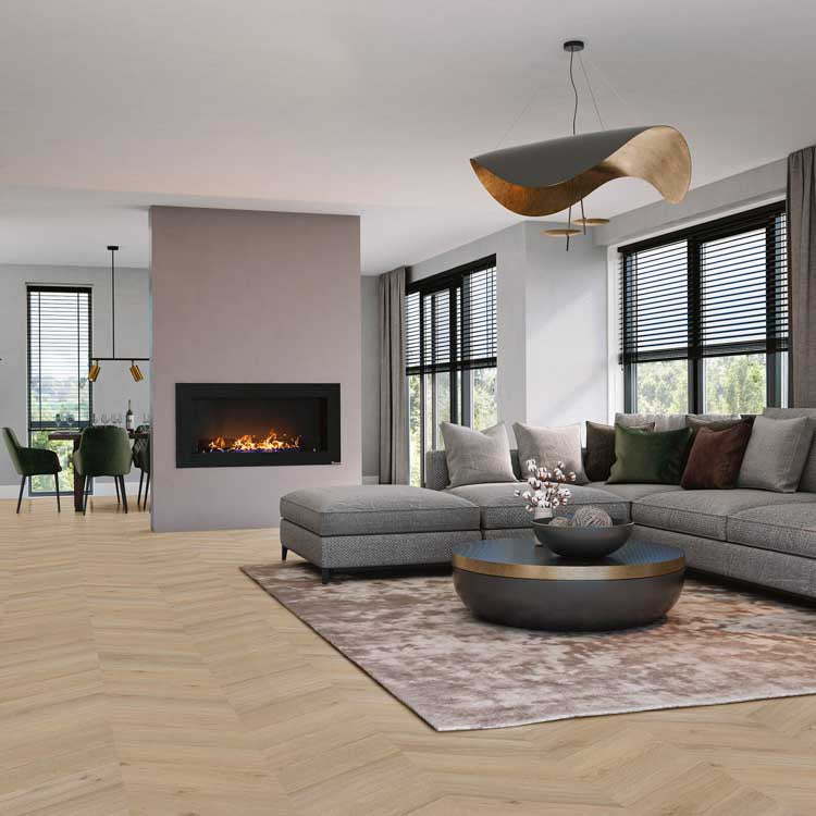 lichte vloer in visgraatpatroon met brede planken in licht eiken kleur, moderne zithoek met grijze hoekbank en ronde zwarte salontafel met gouden rand, taupe kleed, grote ramen met zwarte jaloezieën en open haard in grijze wand