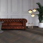 donkergrijze vloer in visgraatpatroon met matte structuur en brede planken naast een cognackleurige leren Chesterfield bank, gouden hanglampen en een grote groene plant in ruimte met lichtgrijze wandpanelen