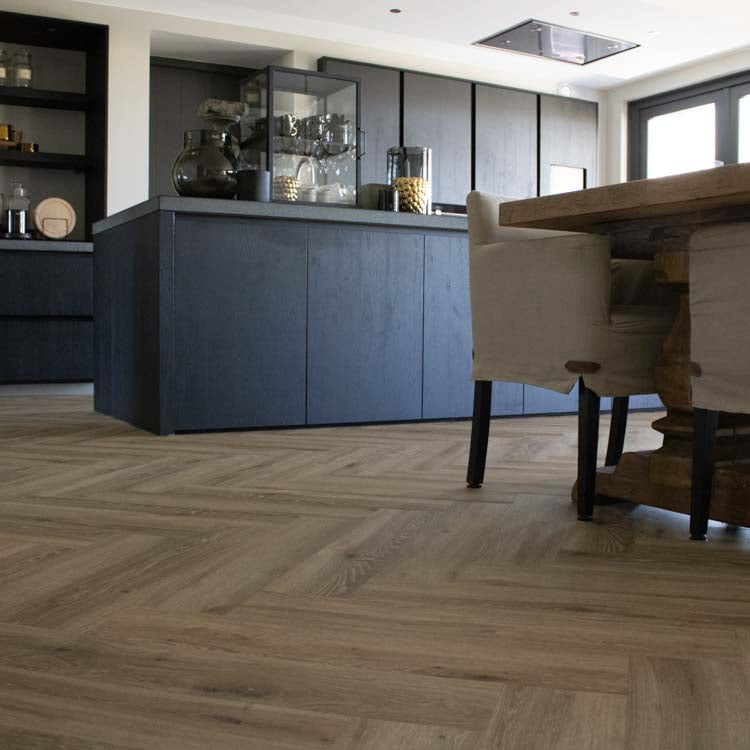Visgraatvloer in licht beige eiken kleur met zichtbare houtnerfstructuur, gecombineerd met een moderne donkere keuken en een eettafel met beige beklede stoelen.