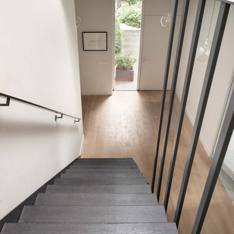 Donkergrijze trap met subtiele houtstructuur en rechte treden, zicht op licht eiken vloer met brede planken en lichte houtnerven in gang met witte muren en glazen balustrade