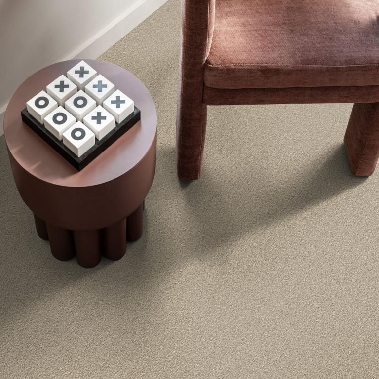 Beige tapijt met fijne, gelijkmatige structuur, gecombineerd met een donkerroodbruine stoffen fauteuil en een rond bruin bijzettafeltje met een noughts-and-crosses spel erop.