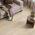 lichte eiken vloer met brede planken en subtiele houtnerfstructuur, gecombineerd met een beige stoffen bank en een kleurrijk geweven plaid met franjes, naast een houten ronde bijzettafel en een vaas met groene takken en paarse bloemen