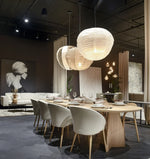 donkergrijze visgraatvloer met matte structuur onder houten eettafel met licht eiken tafelblad en witte gestoffeerde stoelen, warme zachte verlichting door ronde witte papieren hanglampen, modern interieur met beige bank en zwart-wit kunstwerk op achtergrond