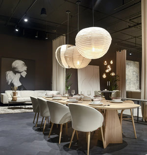 donkergrijze visgraatvloer met matte structuur onder houten eettafel met licht eiken tafelblad en witte gestoffeerde stoelen, warme zachte verlichting door ronde witte papieren hanglampen, modern interieur met beige bank en zwart-wit kunstwerk op achtergrond