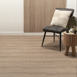 lichte vloer in beige eiken tint met subtiele houtstructuur, strakke stoel met zwart metalen poten en antraciet grijze zitting, decoratief houten bijzettafeltje met planten en kussen met diagonale ribbelstructuur op neutrale achtergrond