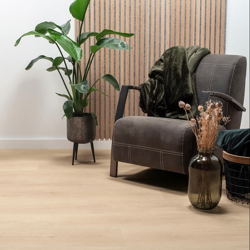 lichte beige vloer met brede planken en subtiele houtnerfstructuur, zachte grijsbruine fauteuil met stiknaden, donkergroen zacht plaid en droogboeket in donkergrijze glazen vaas naast metalen plantenbak met donkergroene grote kamerplant tegen houten lamellenwand
