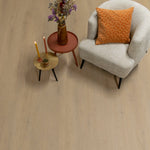 lichte vloer in brede planken met een licht beige eiken houtstructuur, afgebeeld naast een zachtcreme gebouorde fauteuil met een oranje fluwelen kussen, en twee kleine ronde bijzettafels in terracotta en goud met kaarsen en een vaas met droogbloemen erop
