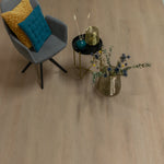 licht beige eiken vloer met zichtbare nerfstructuur en brede planken, sfeervolle zithoek met grijze stoel, blauwe en okergele kussens, en goudkleurige decoratie op zwarte tafeltje