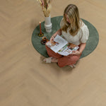 licht beige eiken visgraat vloer met subtiele houtnerfstructuur, brede planken, rustieke matte afwerking onder comfortabele zithoek met groen rond vloerkleed, brandende kaars en gezellige setting met vrouw die tijdschrift bekijkt