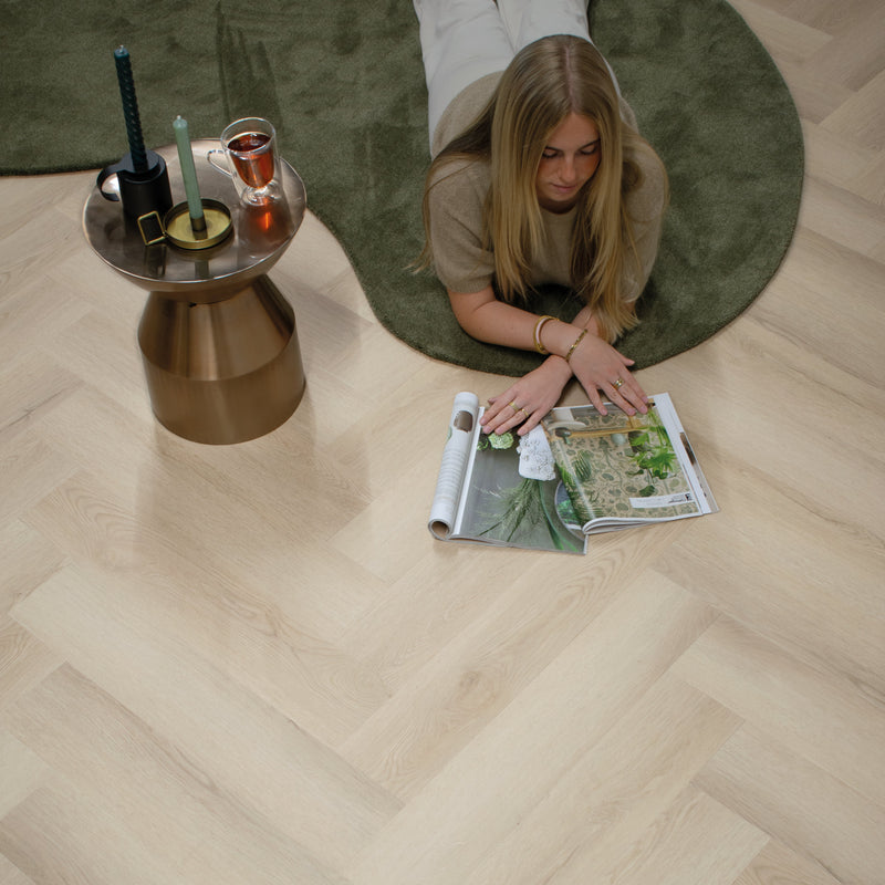 licht eiken vloer met visgraat patroon van brede planken, mat afgewerkt met subtiele houtnerfstructuur, vrouw liggend op rond mosgroen tapijt naast gouden bijzettafel met kaarsen en glas thee