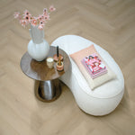 lichte eiken visgraatvloer met subtiele houtnerfstructuur, gecombineerd met een ronde donkere houten bijzettafel, witte decoratieve vaas met roze bloemen, en een zachte witte bouclé fauteuil met roze kussen en stapel boeken erop