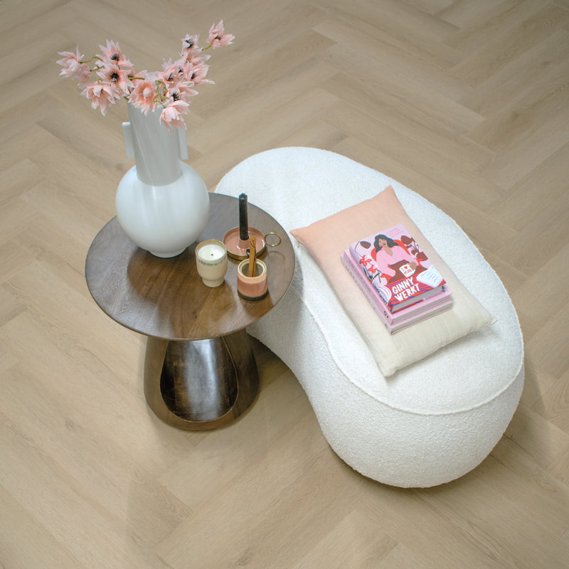 Licht beige eiken vloer met visgraatpatroon en subtiele houtstructuur, gecombineerd met een ronde houten tafel en decoratieve vaas met lichtroze bloemen, witte zachte stoel met kussen en stapel boeken.