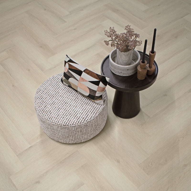 Licht eiken visgraatvloer met subtiele houtnerfstructuur en brede planken, gecombineerd met een ronde poef met grove gebreide structuur en een zwart houten bijzettafeltje met decoratieve vaas en houten kaarsenhouders.