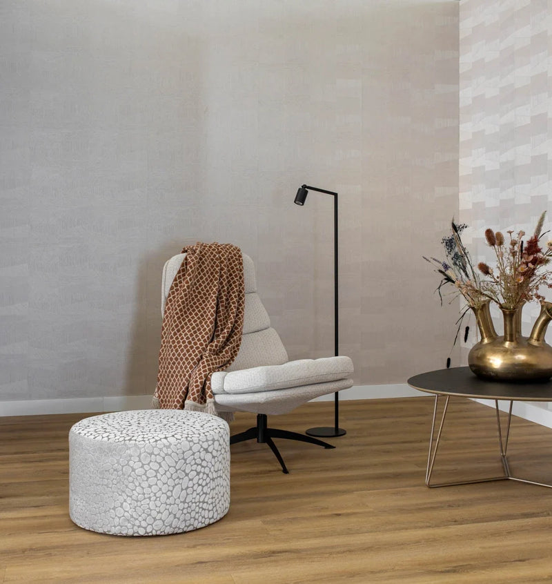 licht eiken vloer met brede planken en een naturel houtstructuur, gecombineerd met een lichtgrijze ronde poef met subtiel kiezels patroon, een beige fauteuil met een bruin geometrisch dessin deken, zwarte vloerlamp en een zwarte salontafel met gouden vaas en droogbloemen in een neutrale woonkamer