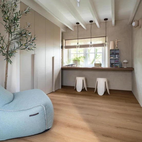 lichte, natuurkleurige houten vloer met brede planken in een rechte patroon, gecombineerd met lichte beige wanden, een lichtblauwe zitzak en houten werkblad met twee witte krukken onder een raam met natuurlijke lichtinval