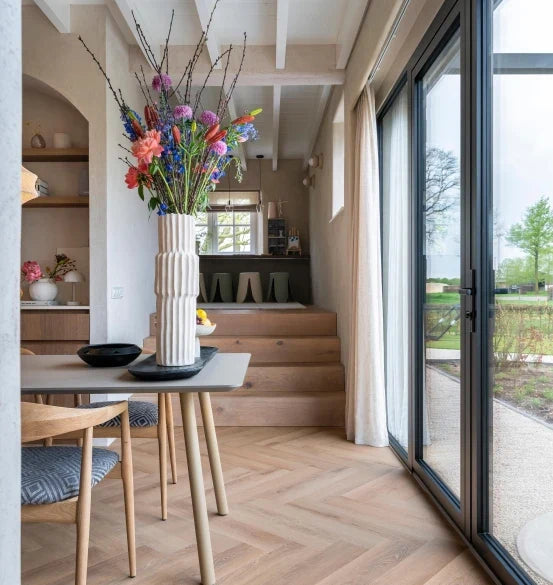 lichte eiken visgraatvloer met brede planken in een moderne eetkamer met grote glazen schuifdeuren, witte muren en een grijs houten tafelblad met een witte vaas met kleurrijke bloemen erop