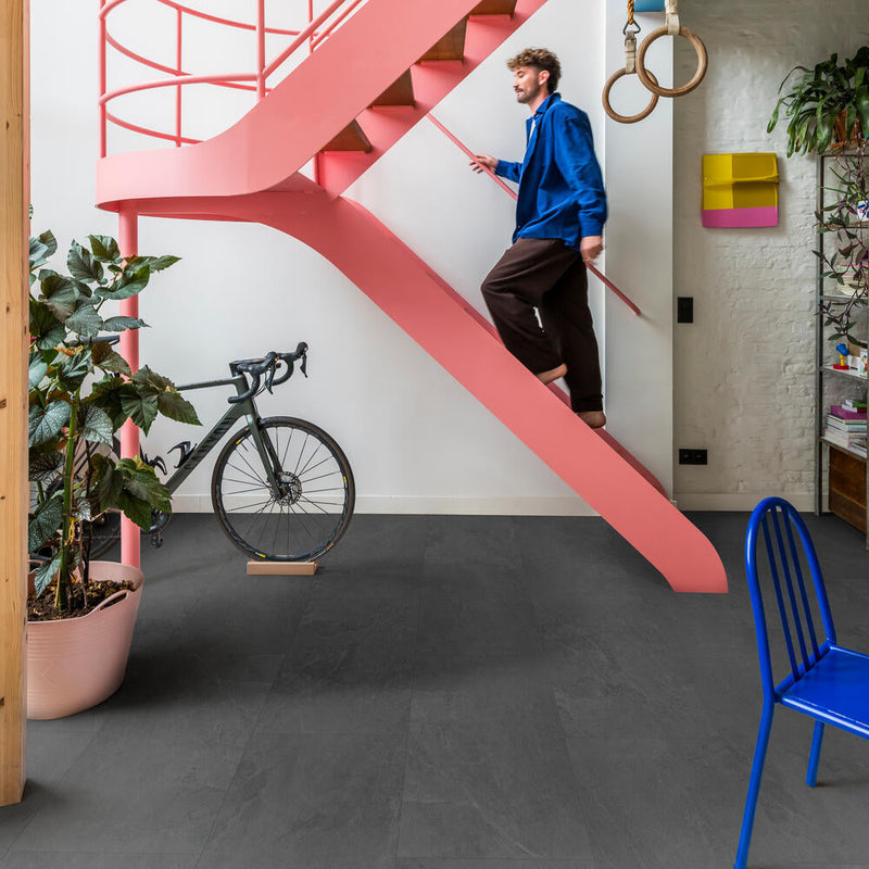 donkergrijze tegelvloer met matte structuur en lichte golvende lijnen, gecombineerd met een felroze spiraalvormige trap en diverse kleurrijke interieuraccenten zoals een blauwe stoel en een groene plant in een lichtroze pot