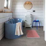 donkergrijze tegelvloer met grote, matte tegels met subtiel structuurpatroon in een badkamer met witte rechthoekige wandtegels, blauwe vrijstaande badkuip en blauw bijzettafeltje