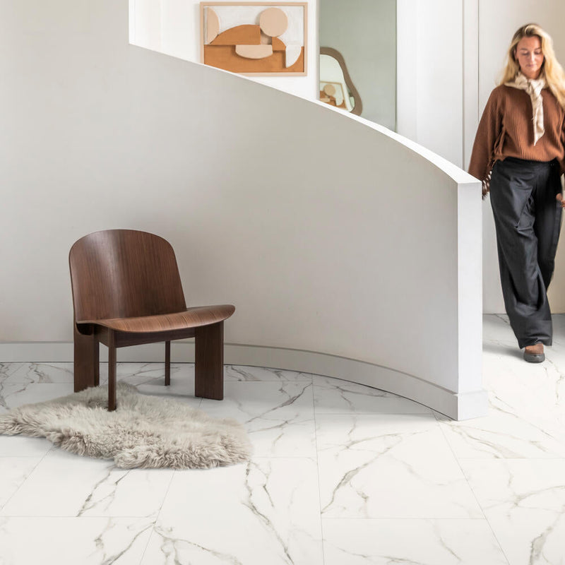 Witte tegelvloer met marmerpatroon in lichte beige en grijsbruine aders, brede tegels, moderne zithoek met donkerbruine houten stoel en lichtgrijze schapenvacht op de vloer.